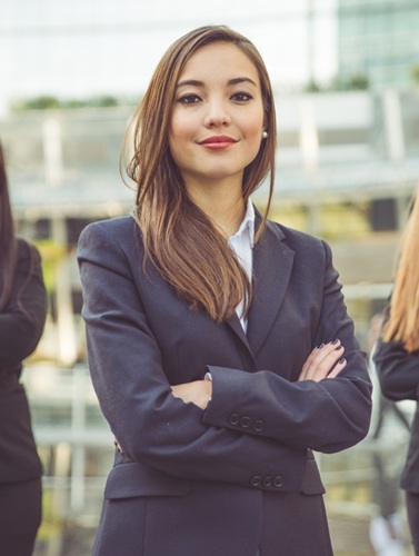 Frau im Business-Anzug steht mit verschränkten Armen, trägt lange braune Haare und blickt selbstbewusst in die Kamera.