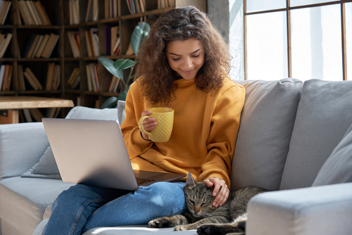 Dunkelhaarige Frau sitzt mit Laptop auf dem Schoss auf der Couch, eine Tasse in der Hand und eine Katze liegt neben ihr und wird von ihr gestreichelt.