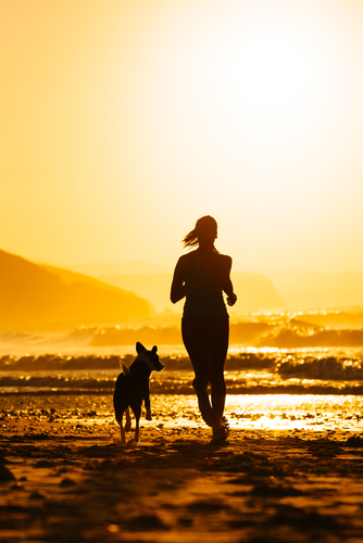 Frau rennt mit einem Hund am Strand entlang, während im Hintergrund Wellen und ein Sonnenuntergang zu sehen sind.