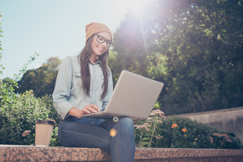 Dunkelhaarige Frau, mit Brille und Mütze, sitzt mit dem Laptop auf dem Schoss im Freien.