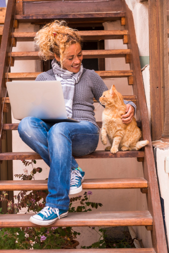 Frau mit lockigem Haar sitzt draußen auf einer Holztreppe, streichelt eine Katze und hält in der anderen Hand einen Laptop.