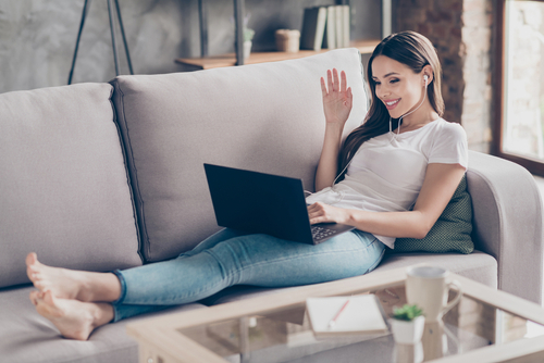 Braunhaarige Frau liegt auf einem Sofa, hat einen Laptop auf dem Schoß und winkt in die Kamera.