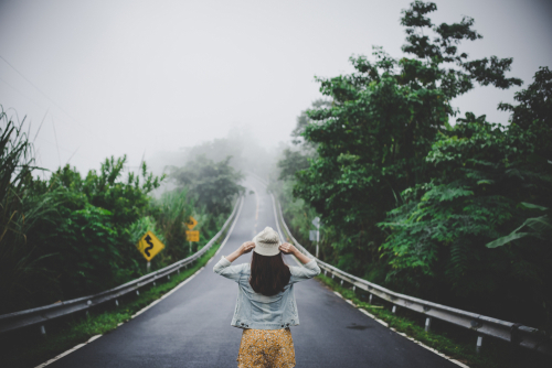 Rückenansicht einer Frau mit Hut, die mittig auf einer Straße steht. Die Straße wird auf beiden Seiten von Bäumen begrenzt.