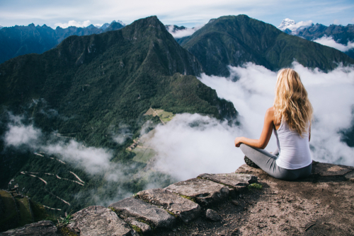 Rückenansicht einer Frau mit langen blonden Haaren, die auf einem Berg meditiert, mit Blick auf Berge und Landschaft.