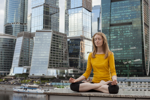 Frau mit langen Haaren meditiert in Lotus-Pose in der Stadt vor Hochhäusern.