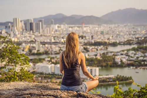 Rückenansicht einer Frau mit langen Haaren, die draußen auf dem Boden meditiert, mit Blick auf die Stadt.
