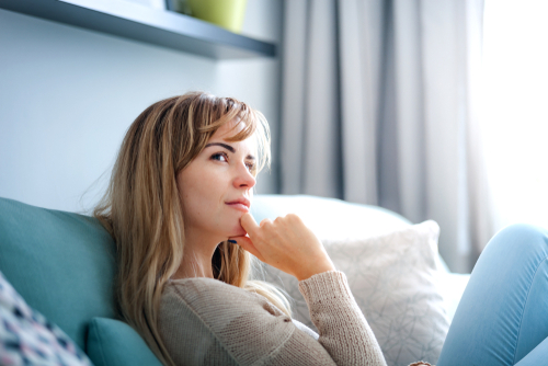 Nachdenkliche Frau mit blonden Haaren auf der Couch, Hand am Kinn.