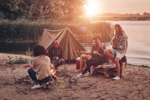 Fünf Freunde sitzen am Lagerfeuer vor einem Zelt am Sandstrand, während die Sonne über dem See untergeht.