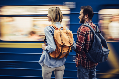 Ein Mann und eine Frau mit Rucksäcken auf den Rücken stehen nebeneinander am Bahnsteig und schauen auf einen vorbeifahrenden Zug.